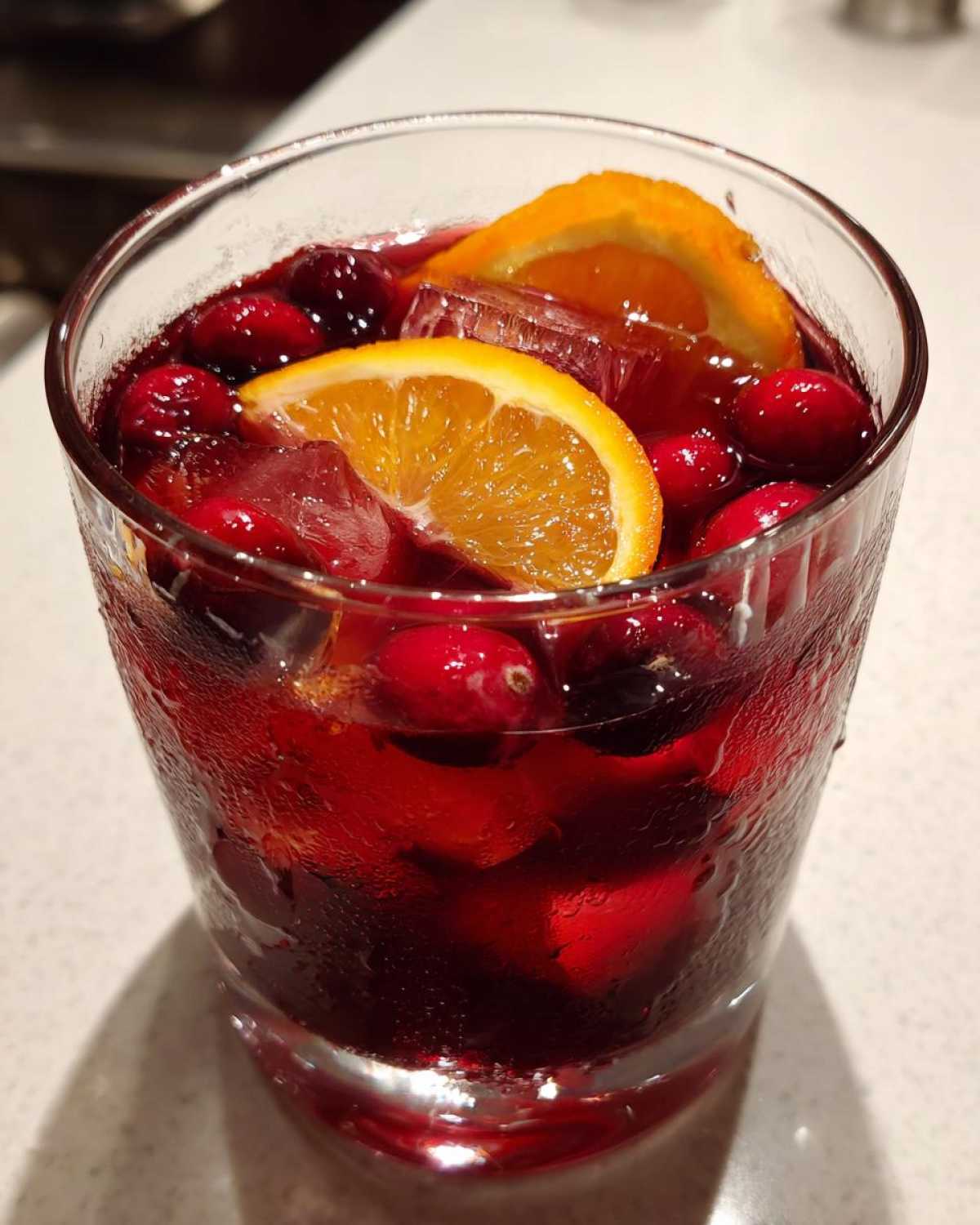 Close-up of a refreshing glass of Thanksgiving punch, filled with deep red liquid, ice, whole cranberries, and orange slices.