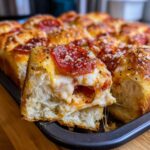 A close-up of one gooey piece being pulled from a pan of baked pizza sliders topped with pepperoni and cheese.