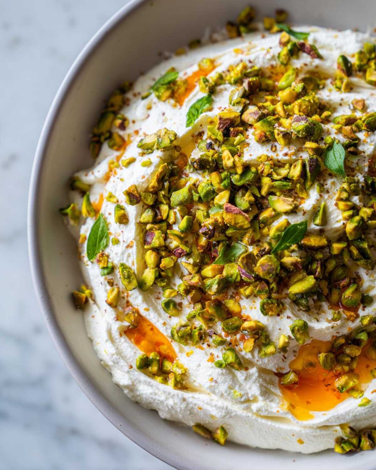 Close-up of a creamy ricotta dip topped generously with chopped green pistachios, drizzled with orange-colored honey, and garnished with mint leaves.