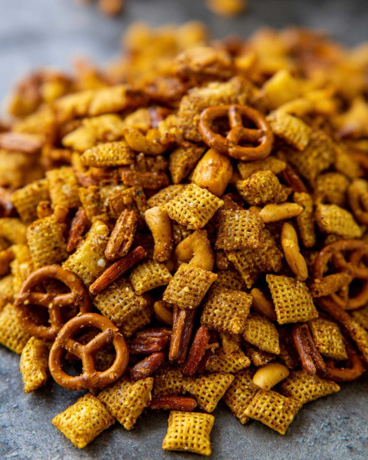 A close-up, macro shot of a pile of seasoned chex party mix featuring square cereal pieces, mini pretzels, and nuts.