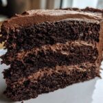 Close-up of a moist slice of homemade chocolate cake with three layers and thick chocolate frosting.