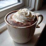 A mug of rich homemade hot cocoa topped with a swirl of whipped cream, sitting by a window.