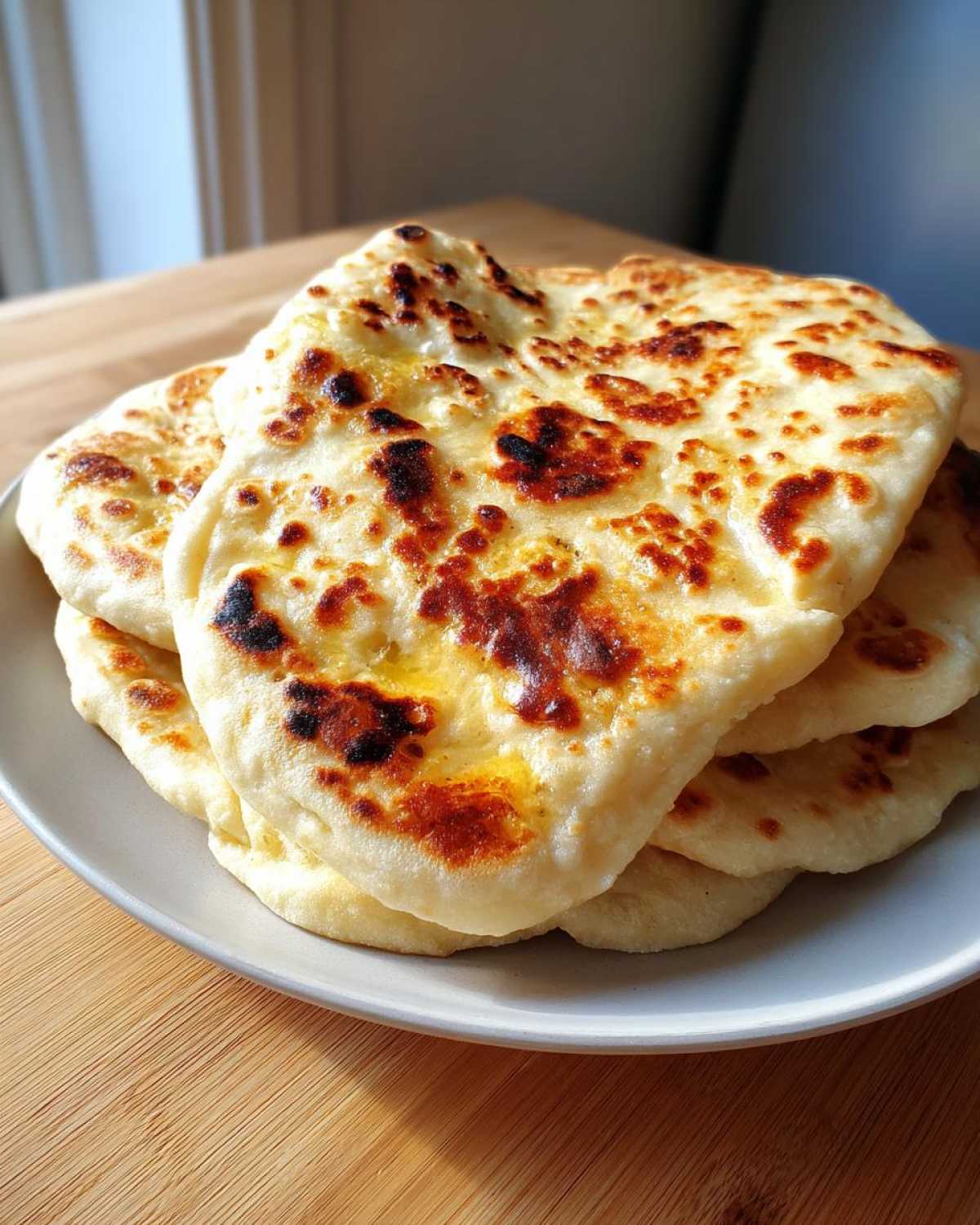 A stack of freshly cooked, golden-brown naan bread with charred spots, resting on a light gray plate.