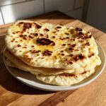 A stack of freshly cooked, buttery naan bread with charred spots, resting on a plate.