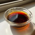 Close-up of rich, dark homemade teriyaki sauce in a clear glass bowl, glistening in sunlight.