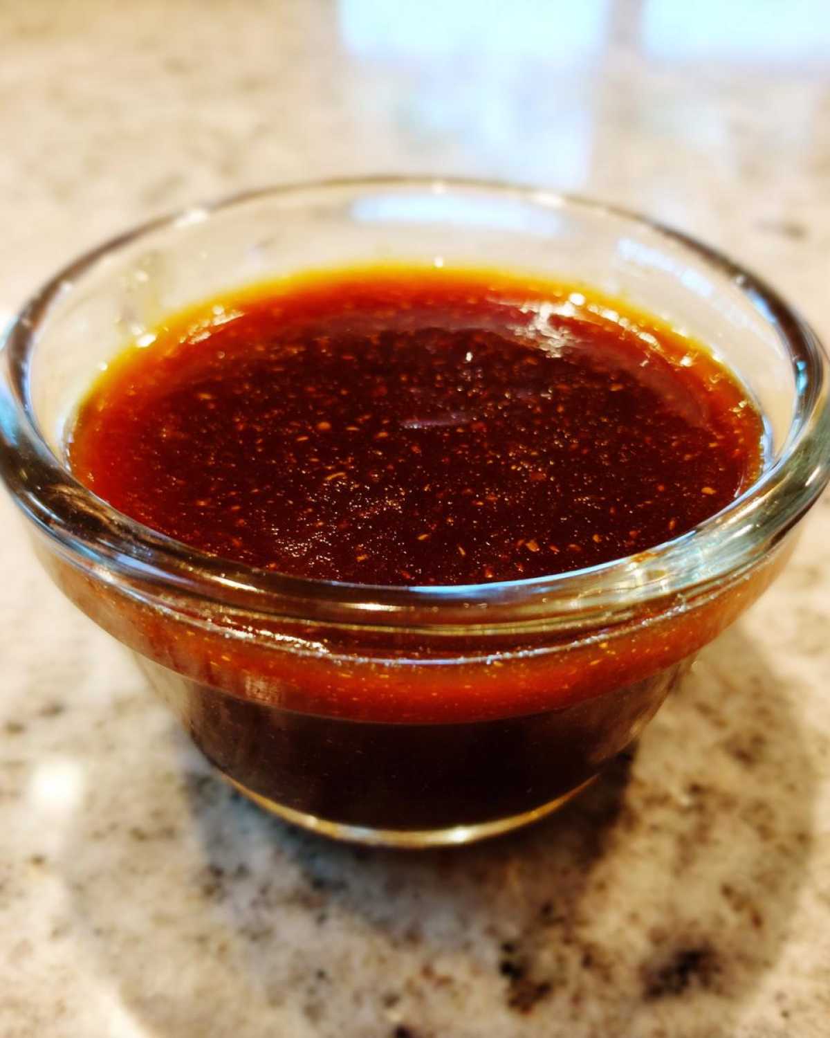 Close-up of rich, dark homemade teriyaki sauce with visible spices in a small clear dipping bowl.