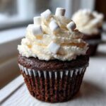 Close-up of a dark chocolate hot chocolate cupcake topped with white frosting and mini marshmallows.