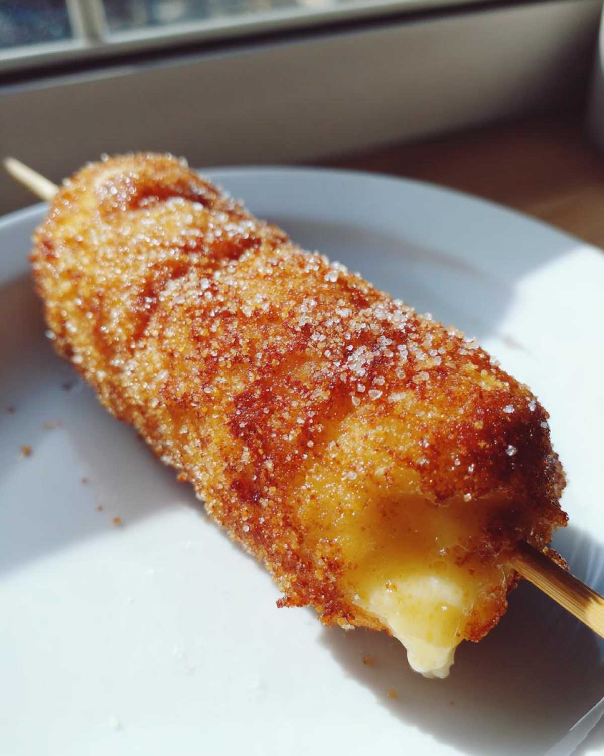 Close-up of a freshly fried korean corn dog on a stick, coated in cinnamon sugar with melted cheese oozing out.