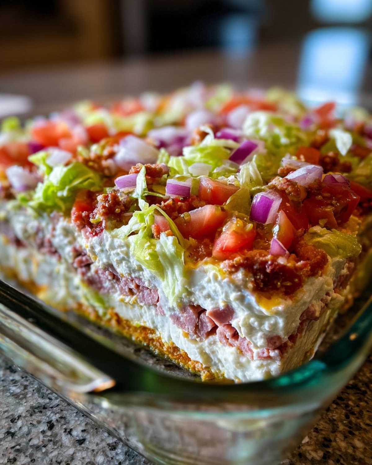 Close-up of a thick, layered hoagie dip topped with lettuce, tomatoes, and red onions in a glass dish.