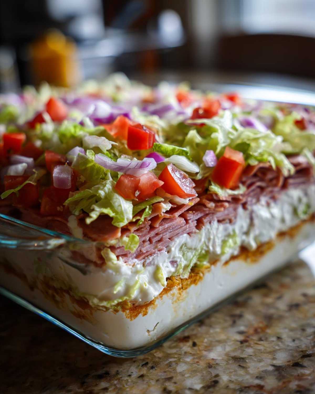 Close-up of a layered hoagie dip in a glass baking dish, topped with lettuce, tomatoes, and onions.