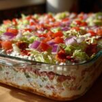 Close-up of a layered hoagie dip topped with lettuce, tomatoes, red onion, and pepperoni.