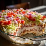 Cross-section view of a layered hoagie dip showing cream cheese, meat, and cheese layers topped with shredded lettuce, tomatoes, and red onion.