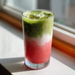 A tall glass showing a layered strawberry matcha drink with a bright red bottom layer, a white middle layer, and a foamy green matcha top layer with ice.