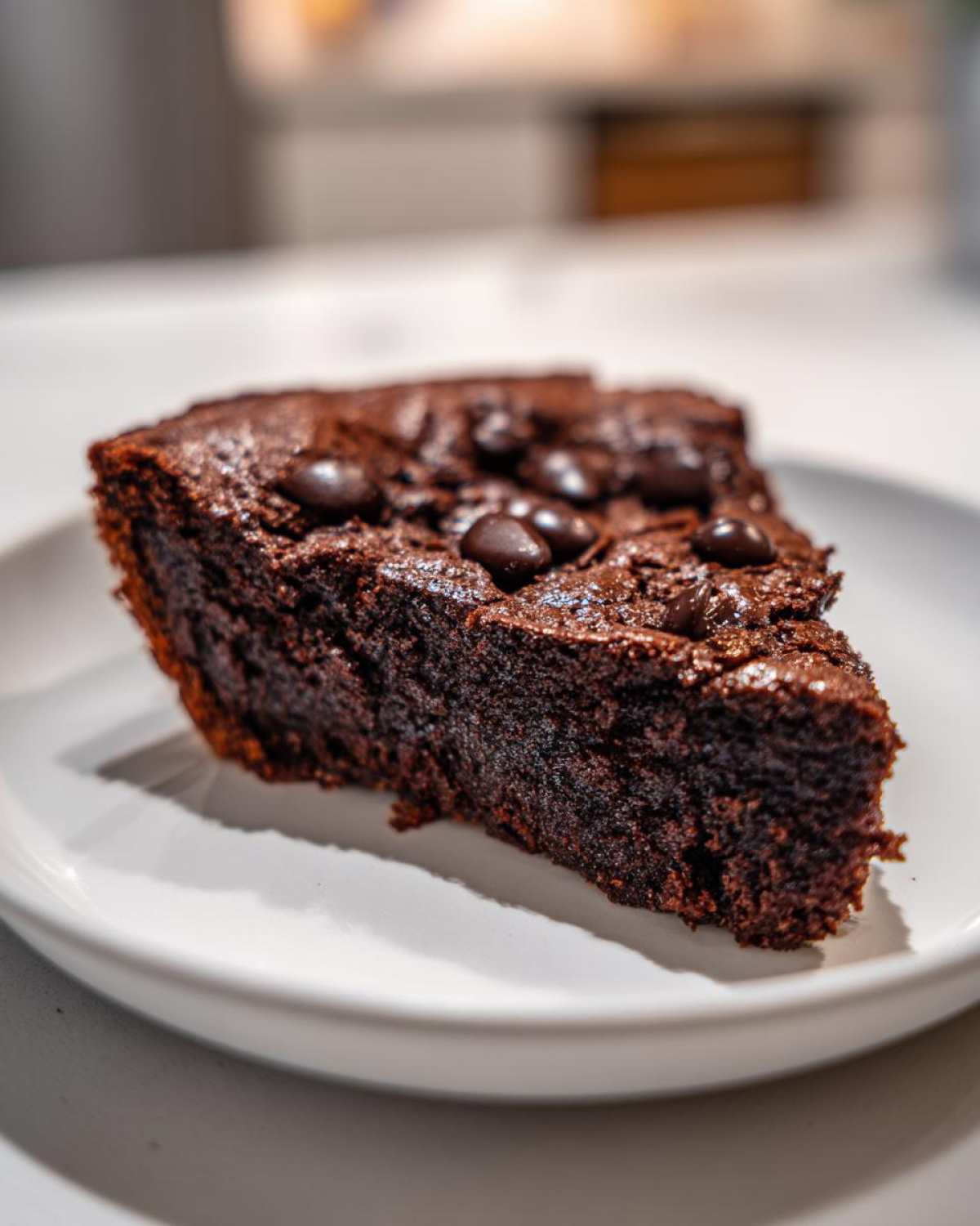 A close-up of a dark, moist slice of fudge pie topped with chocolate chips, sitting on a white plate.