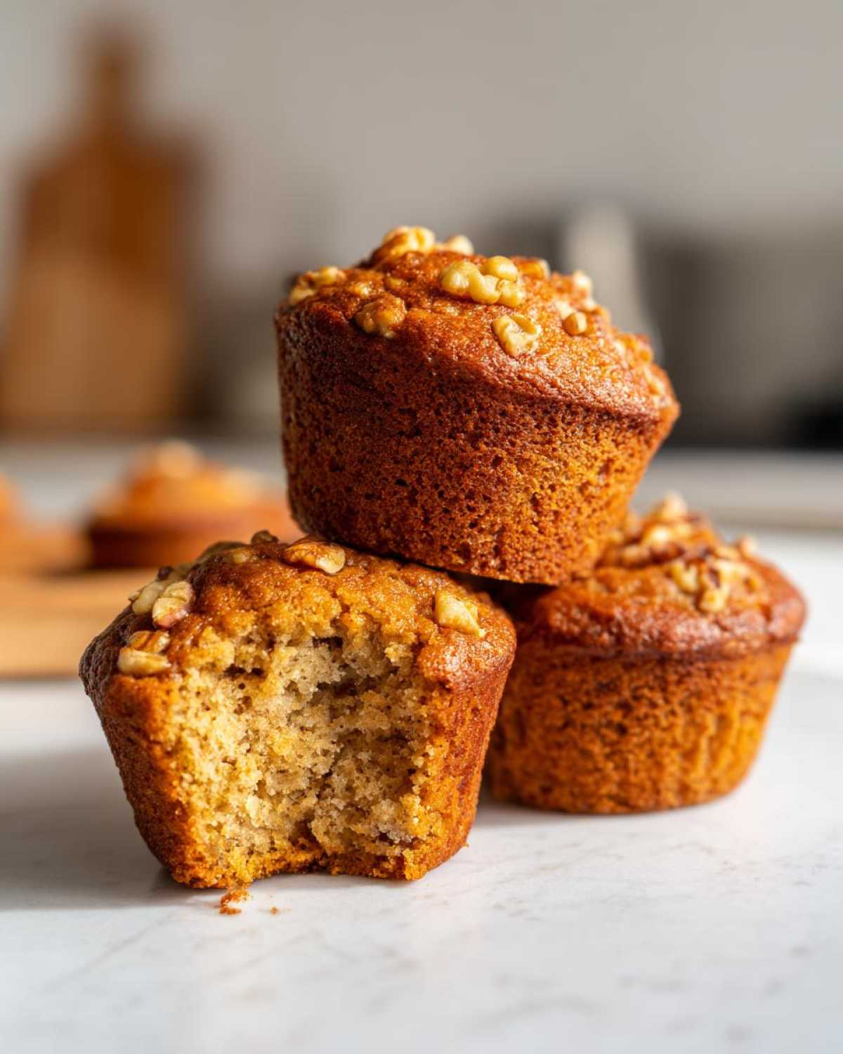 A stack of three moist banana nut muffins, one with a bite taken out, topped with walnuts.