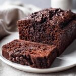 A moist, dark chocolate bread loaf, partially sliced, topped with chocolate chips on a white plate.
