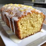 Close-up of a freshly baked lemon loaf cake, drizzled with white icing and topped with bright yellow lemon zest.