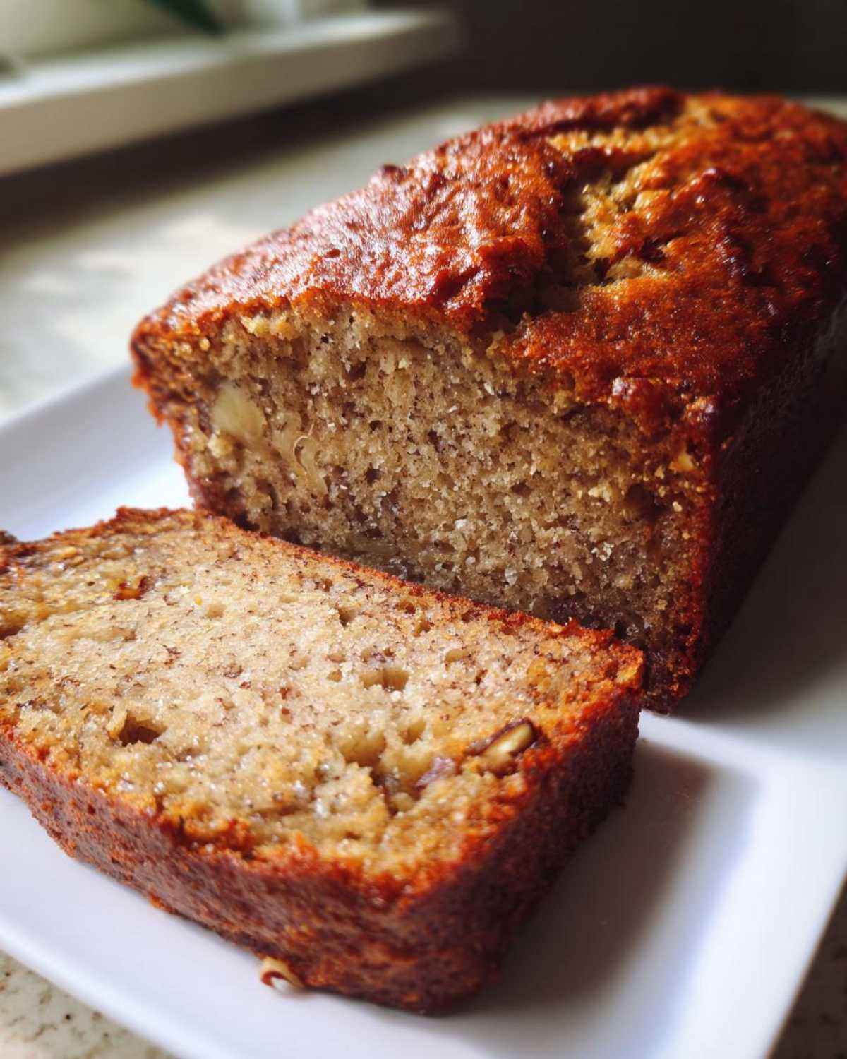 A moist loaf of sugar free banana bread, with one thick slice cut and resting beside it on a white plate.