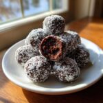A stack of rich, dark bourbon balls coated in shredded coconut, with one cut open showing the fudgy interior.