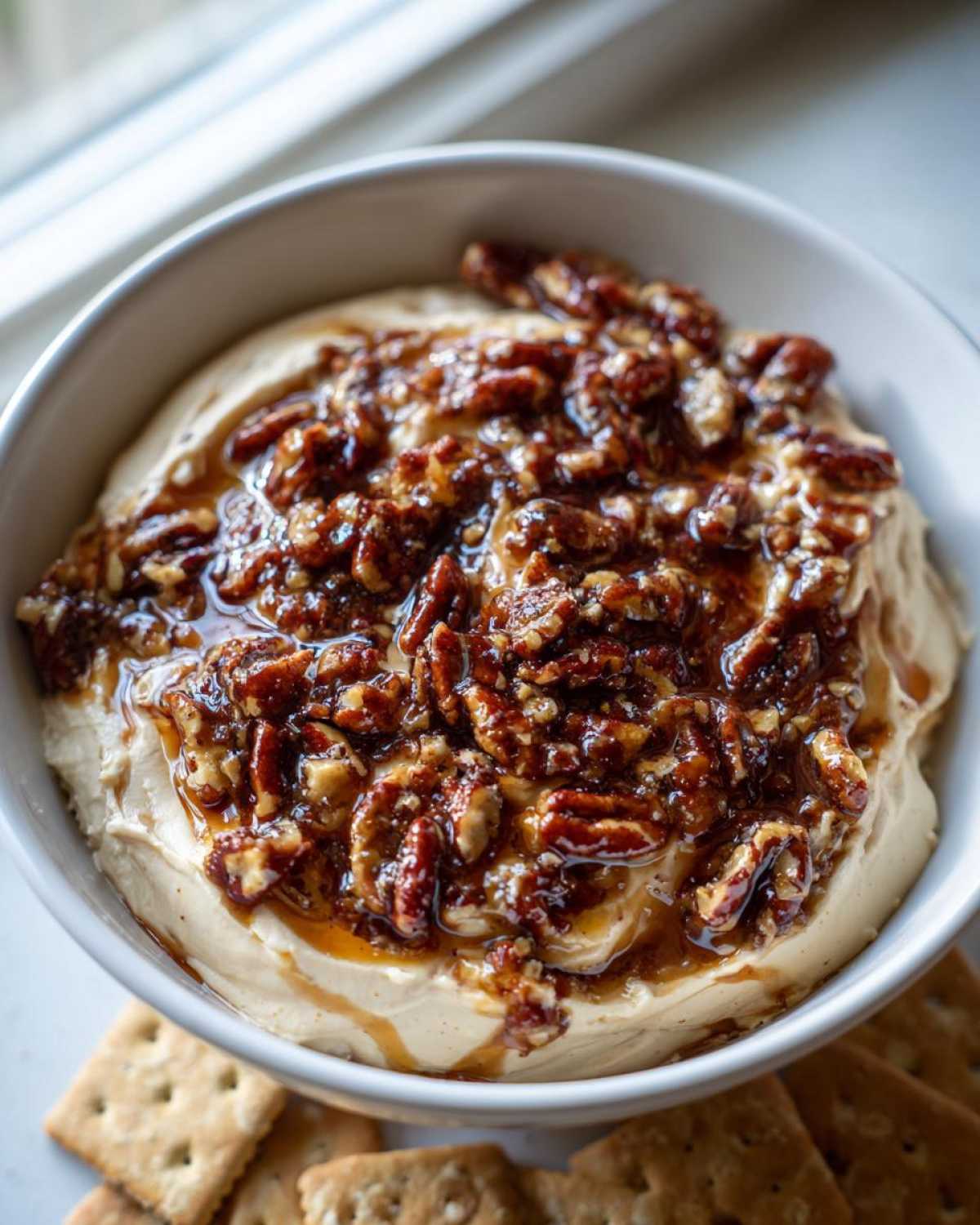 Close-up of a creamy no-bake pecan pie dip topped generously with glazed pecans and caramel sauce, served with crackers.