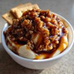 Close-up of a creamy no-bake pecan pie dip topped with glossy, syrupy pecans, served with crackers in the background.