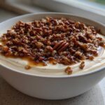Close-up of a creamy, light-colored pecan pie dip topped generously with glazed, chopped pecans and caramel sauce.