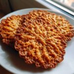 Three golden brown, lacy oatmeal lace cookies with delicate, crispy edges resting on a white plate near a window.
