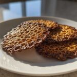 Four golden brown, delicate oatmeal lace cookies with a honeycomb texture stacked on a white plate.