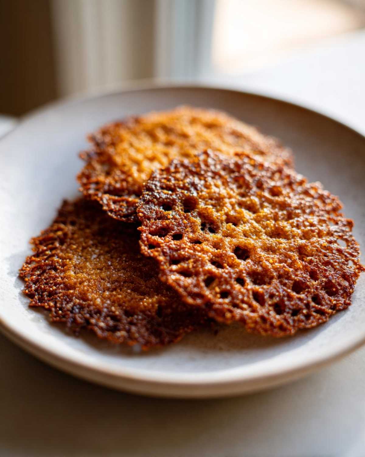 Close-up of three dark golden, lacy oatmeal lace cookies stacked on a light ceramic plate.
