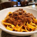 Close-up of rich, meaty bolognese sauce generously spooned over fresh tagliatelle pasta in a white bowl.