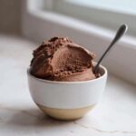 A rich, dark scoop of chocolate mousse served in a small white and tan ceramic bowl with a spoon.