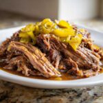 Close-up of tender, shredded Mississippi pot roast served in its juices, topped with yellow pepperoncini slices.