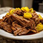 A close-up of tender, shredded Mississippi pot roast topped with bright yellow pepperoncini slices on a white plate.