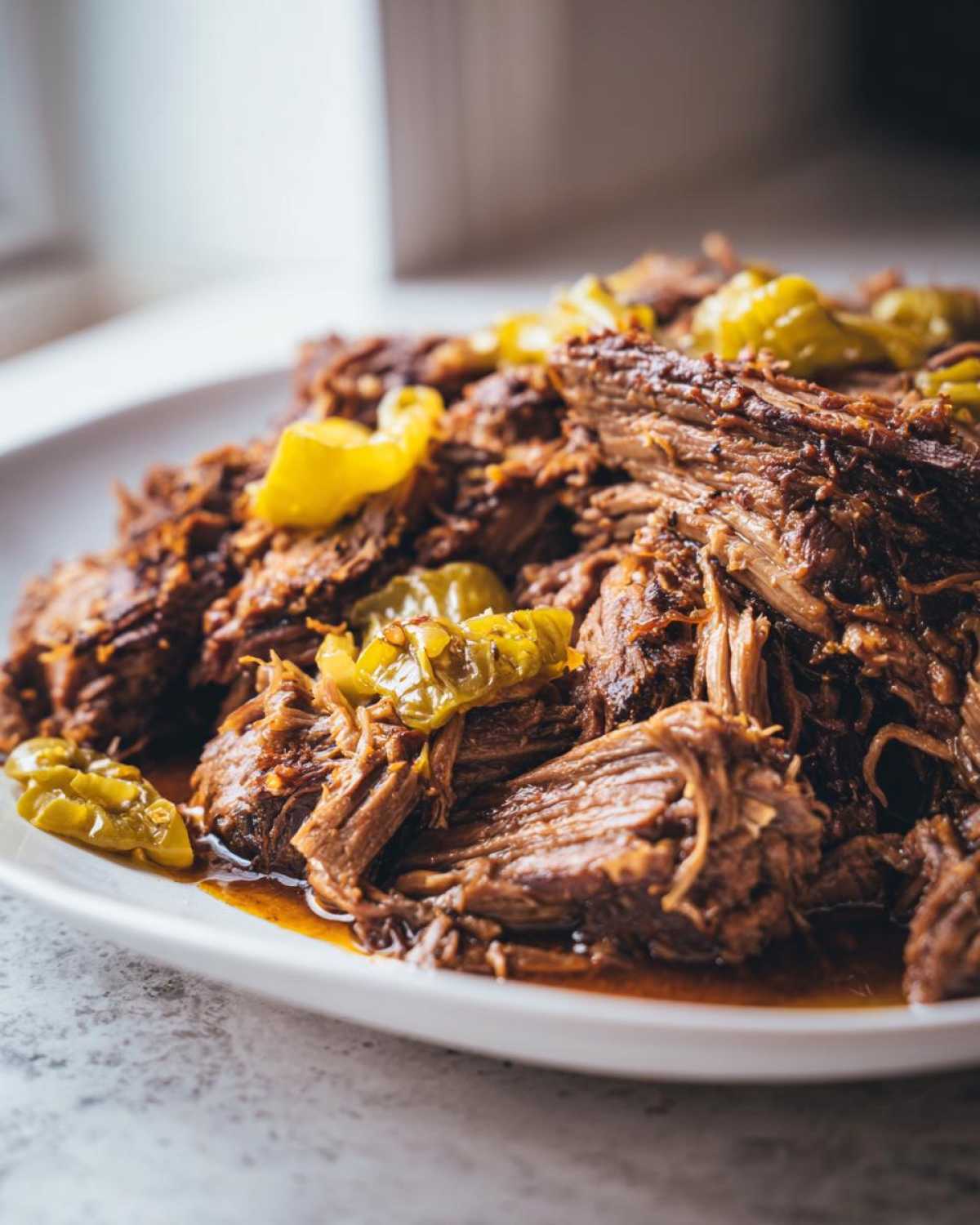 Close-up of shredded, juicy Mississippi pot roast topped with bright yellow pepperoncini peppers on a white platter.
