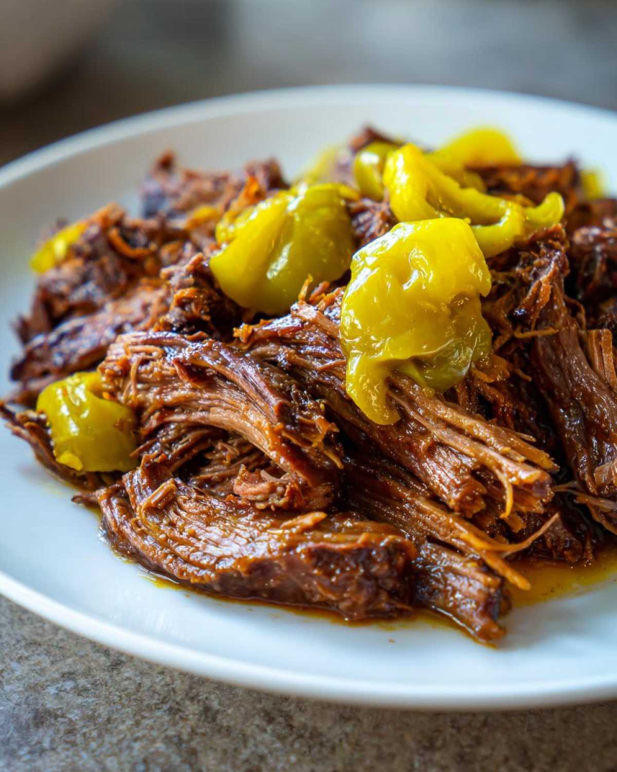 Close-up of tender, shredded Mississippi pot roast topped with bright yellow pepperoncini peppers on a white plate.