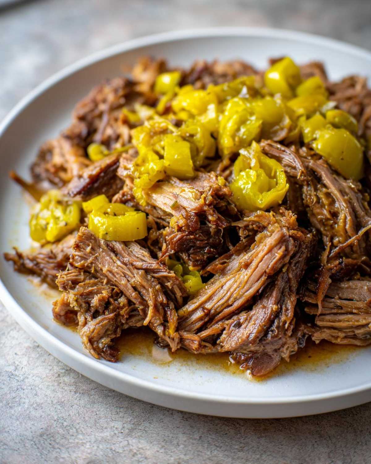 Close-up of shredded Mississippi pot roast topped with bright yellow pepperoncini peppers on a white plate.
