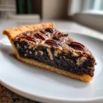 A close-up of a perfect slice of chocolate pecan pie with a flaky crust and whole pecans on top.