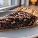 A close-up of a rich, dark slice of chocolate pecan pie with a flaky crust on a white plate.