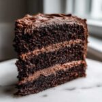 Close-up of a moist, three-layer slice of homemade chocolate cake with thick chocolate frosting.