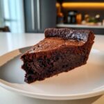 A decadent slice of hot fudge pie with a dense, fudgy interior and slightly cracked top, served on a white plate.