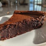 A close-up slice of dense, dark chocolate hot fudge pie resting on a white plate.