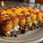 A close-up slice of baked tater tot casserole showing crispy tots, melted cheese, and a ground beef layer.