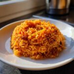 A close-up, sunlit image of a perfectly molded mound of vibrant orange smoky jollof rice on a white plate.