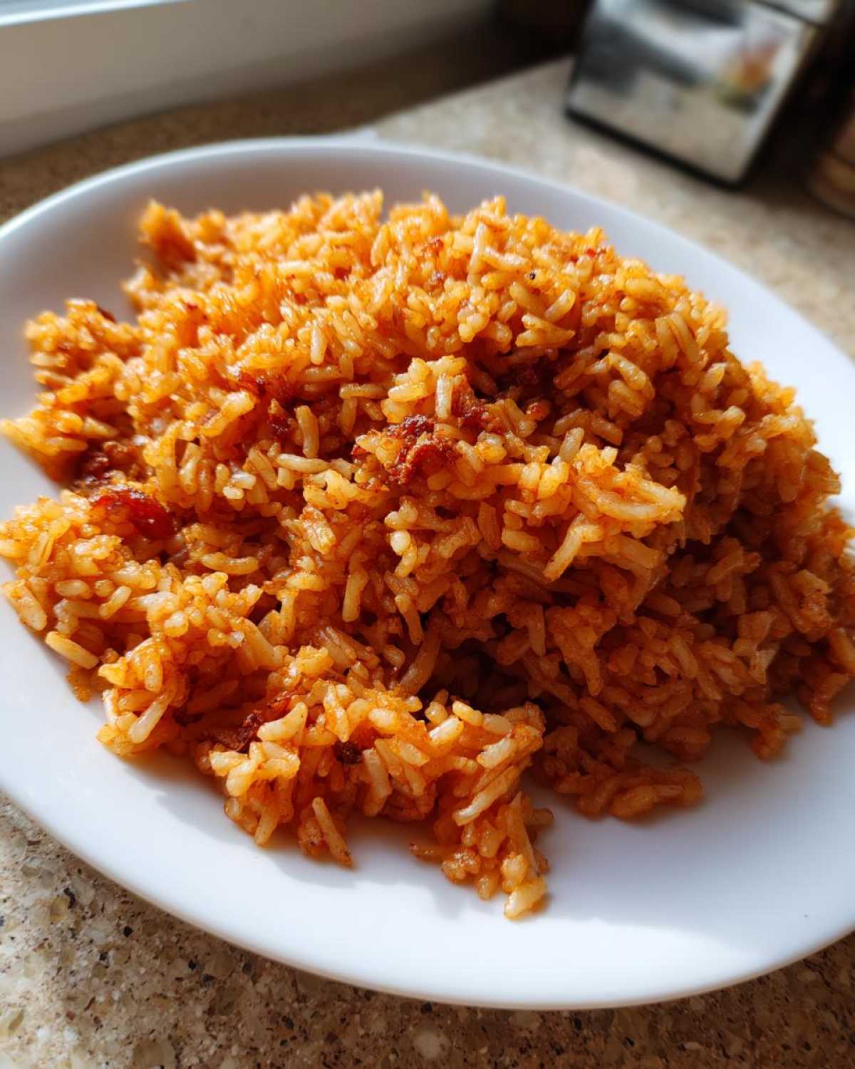 A close-up of fluffy, reddish-orange jollof rice served on a white plate, showing its smoky texture.