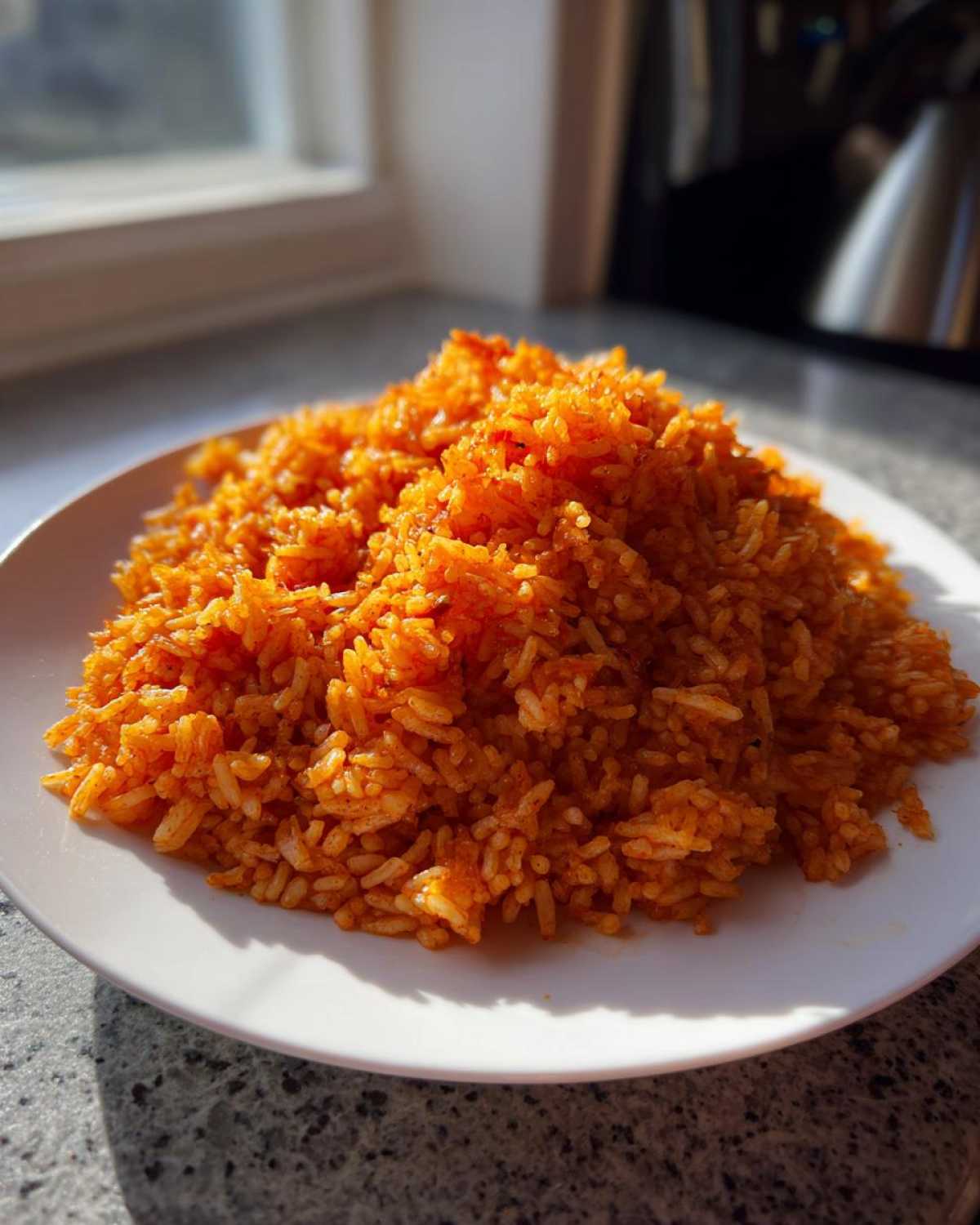 A mound of vibrant, orange-red smoky jollof rice served on a white plate, illuminated by natural light.