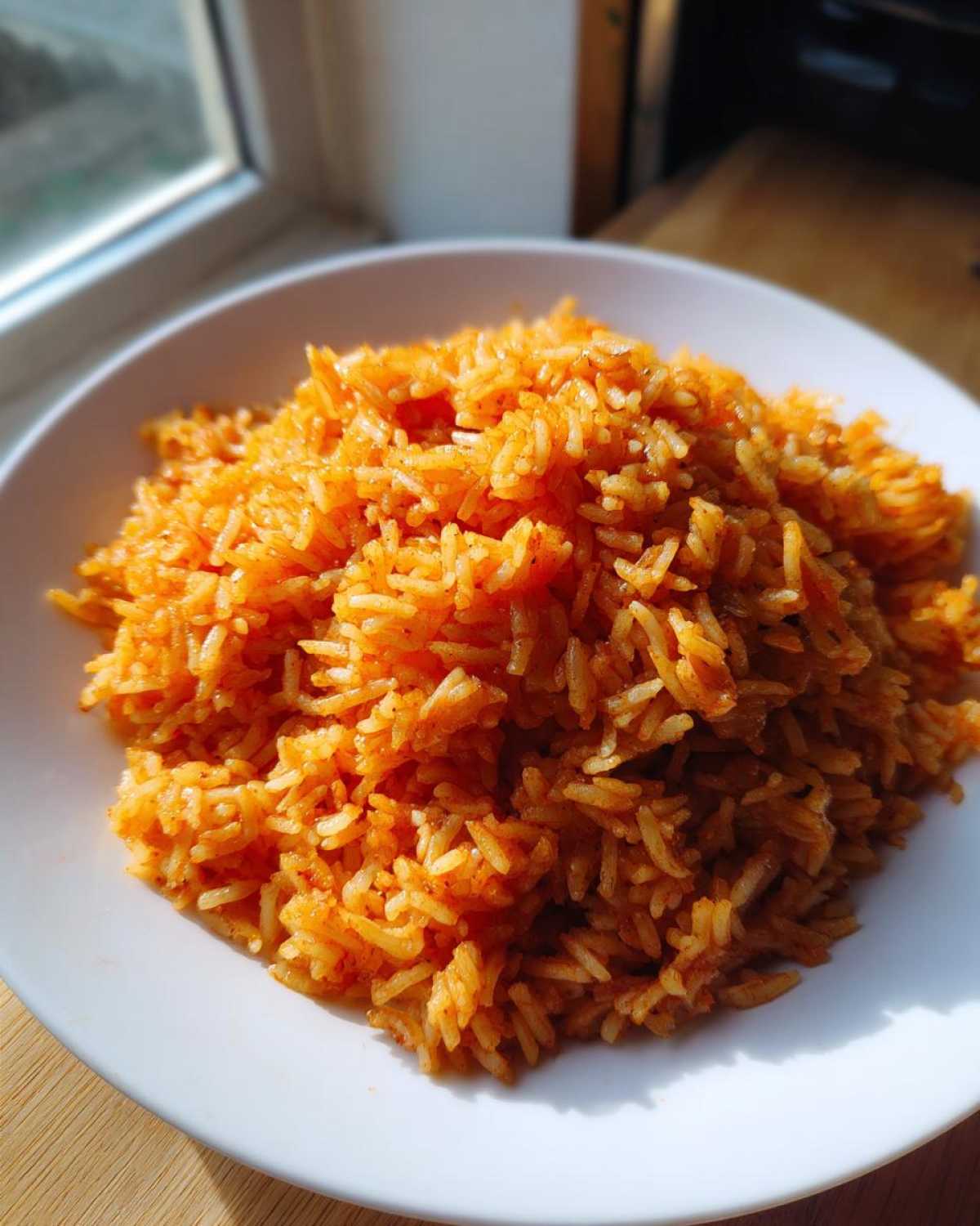 A close-up of perfectly cooked, vibrant orange smoky jollof rice piled high on a white plate, backlit by a window.