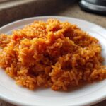 Close-up of vibrant, reddish-orange Smoky jollof rice piled high on a white plate.