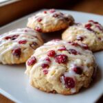 Four soft cranberry orange cookies topped with white glaze and dried cranberries on a white plate.