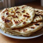 A stack of freshly made, soft naan bread recipe rounds, brushed with melted butter and showing charred spots.