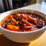 Close-up of a steaming white bowl filled with rich, hearty pumpkin chili featuring ground meat, kidney beans, and chunks of orange pumpkin.